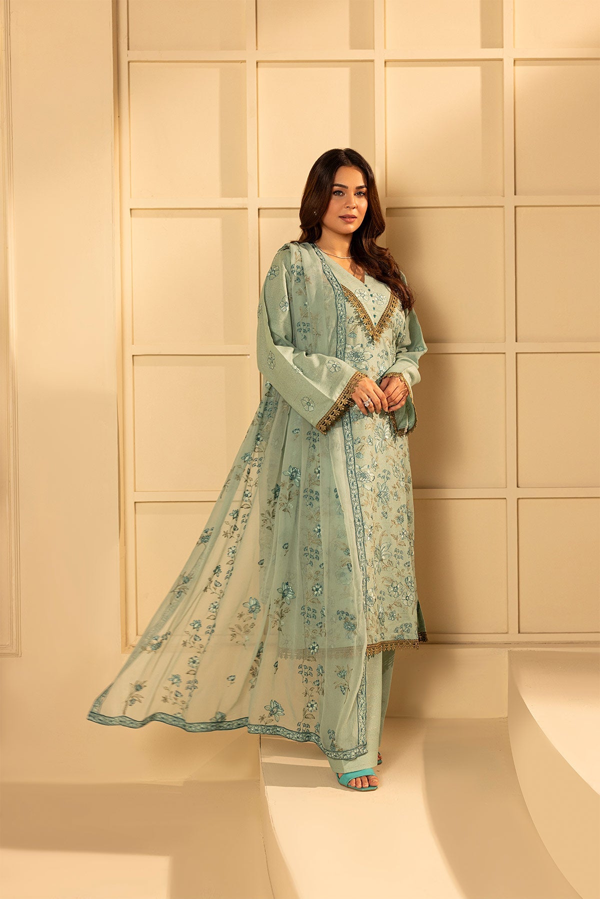 Woman in a light blue traditional outfit standing against a beige wall.