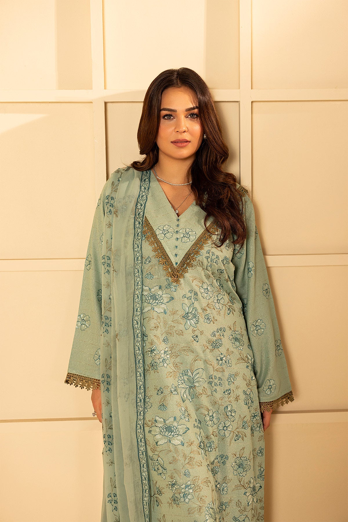 Woman wearing a light green traditional outfit with floral patterns against a beige wall.